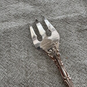 K&ouml;nnte beinhalten: Eine silberfarbene Metallgabel mit drei Zinken und einer texturierten, geb&uuml;rsteten Oberfl&auml;che im mittleren Teil des Kopfes. Der Griff und die Basis der Zinken weisen ein gepr&auml;gtes, erhabenes Schn&ouml;rkelmuster auf, das sich den Stiel entlangzieht.