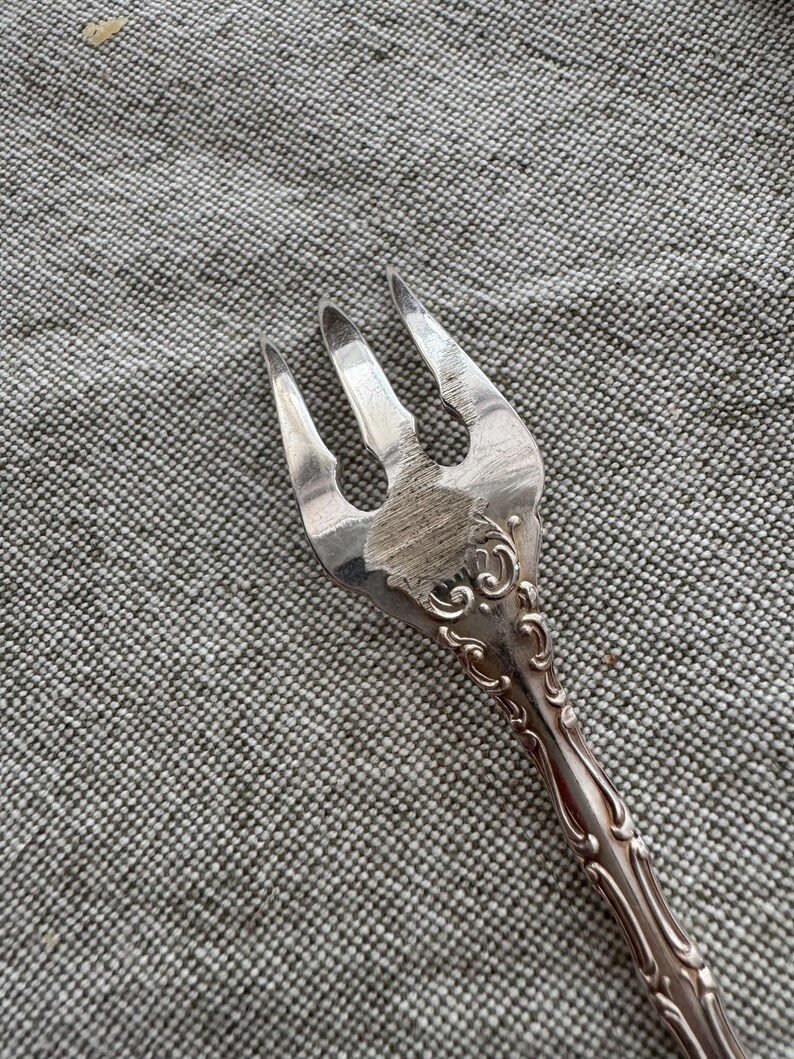 K&ouml;nnte beinhalten: Eine silberfarbene Metallgabel mit drei Zinken und einer texturierten, geb&uuml;rsteten Oberfl&auml;che im mittleren Teil des Kopfes. Der Griff und die Basis der Zinken weisen ein gepr&auml;gtes, erhabenes Schn&ouml;rkelmuster auf, das sich den Stiel entlangzieht.