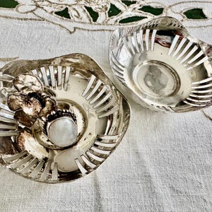 May include: Two silver-toned, openwork bowls. One bowl features a floral design and a central, round, white stone. The other bowl is a simple, openwork design. Both bowls are on a white, textured surface.