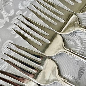 May include: Close-up of several silver forks with tines pointing upwards. The forks are arranged diagonally across a light gray patterned surface. The silverware has a shiny, reflective quality.