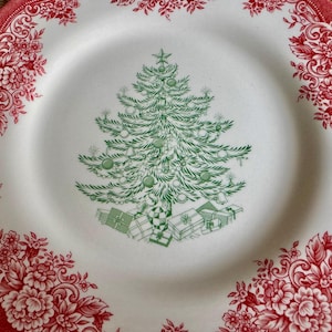 May include: White ceramic plate with a red floral border. The center features a green Christmas tree design with ornaments and gifts. A festive holiday plate for serving food.