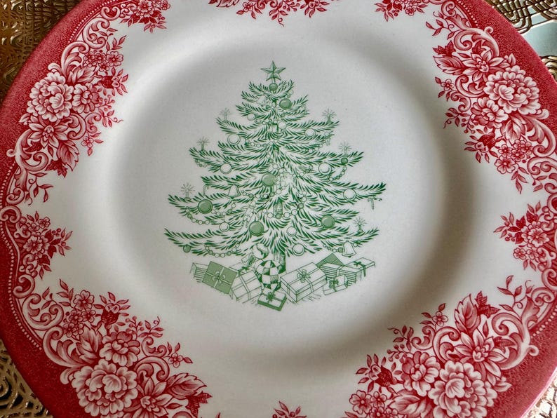 May include: White ceramic plate with a red floral border. The center features a green Christmas tree design with ornaments and gifts. A festive holiday plate for serving food.