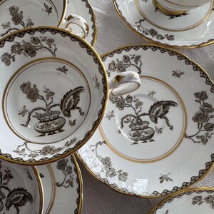 May include: A set of five white teacups and saucers with black and gold floral designs. The cups and saucers have a delicate, intricate pattern.
