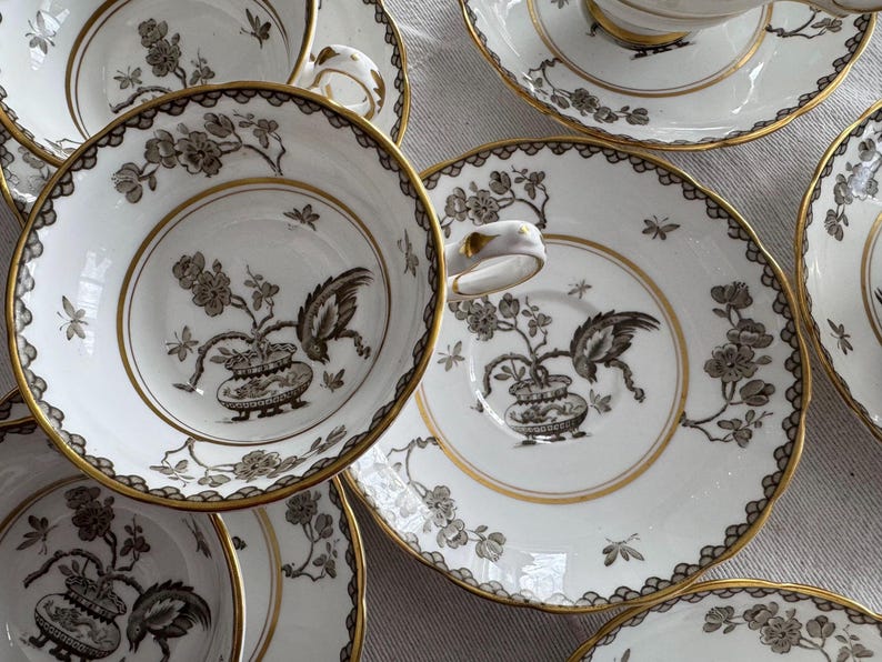 May include: A set of five white teacups and saucers with black and gold floral designs. The cups and saucers have a delicate, intricate pattern.