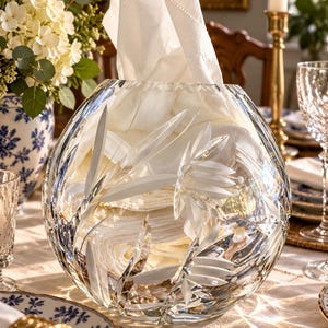 May include: A clear, round crystal bowl featuring etched leaf and floral patterns on its exterior. The bowl is filled with folded white linen napkins, with one napkin extending upward from the center of the vessel.