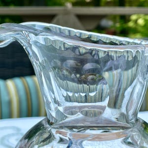 May include: Close-up of a clear glass creamer with a decorative rim and handle. The creamer has a rounded base and a spout for pouring. The glass is transparent, allowing light to pass through, and is set against a blurred background.