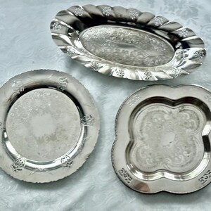 May include: Three silver-colored serving trays. One is oval with a scalloped edge, another is round with a decorative border, and the third is a four-lobed shape with an ornate design. All have a reflective surface.