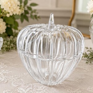 May include: A clear glass pumpkin-shaped container with a removable lid and a pointed stem handle. The body features vertical ribbed detailing throughout, creating a segmented appearance. The glass is transparent and smooth with a reflective surface.