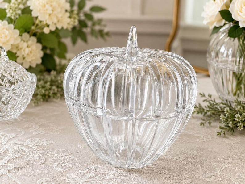 May include: A clear glass pumpkin-shaped container with a removable lid and a pointed stem handle. The body features vertical ribbed detailing throughout, creating a segmented appearance. The glass is transparent and smooth with a reflective surface.