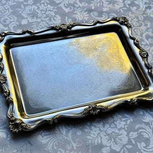 Puede incluir: Una bandeja de servicio rectangular de metal plateado con un borde elevado y ornamentado que presenta motivos en forma de concha en las esquinas y los bordes. El centro de la bandeja muestra un sutil patr&oacute;n floral grabado y pulido.