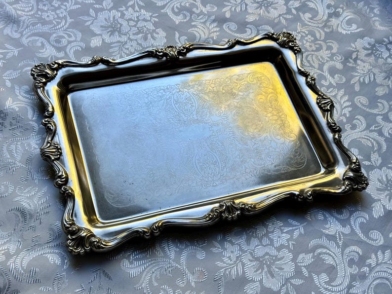 Puede incluir: Una bandeja de servicio rectangular de metal plateado con un borde elevado y ornamentado que presenta motivos en forma de concha en las esquinas y los bordes. El centro de la bandeja muestra un sutil patr&oacute;n floral grabado y pulido.