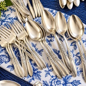 May include: A collection of silver-toned metal flatware including several forks and spoons with ornate floral detailing on the handles. The utensils are arranged on a white cloth featuring a blue floral pattern, with additional pieces visible.
