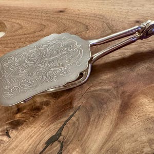 May include: Silver-plated serving tongs with a decorative, ornate design. The tongs have a long handle and a flat, oval-shaped head.