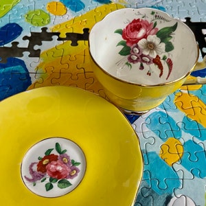 Pu&ograve; includere: Un set composto da tazza e piattino giallo brillante con interno e centro bianchi decorati da un bouquet di fiori colorati. La tazza presenta un bordo smerlato e un manico con finiture dorate, mentre il piattino ha un bordo smerlato abbinato e un motivo floreale centrale.