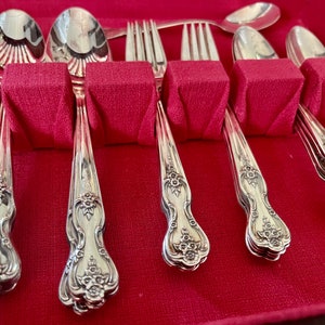 May include: A set of silver-toned flatware including spoons and forks arranged in a red velvet-lined storage case. Each piece features an ornate floral embossed pattern on the handle, with polished bowls and tines reflecting light clearly.