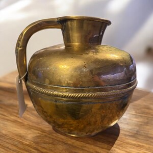 K&ouml;nnte beinhalten: Ein antiker Messingkrug mit rundem Korpus und einem einzelnen Henkel. Der Krug hat ein dekoratives Band um die Oberseite.