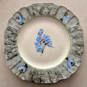 Op de afbeelding: Een ronde keramische bord met een geschulpte rand en een cr&egrave;mekleurig middenstuk beschilderd met een blauwe bloem. De brede rand heeft een getextureerde, gemarmerde grijze afwerking met vier handgeschilderde blauwe bloemen.