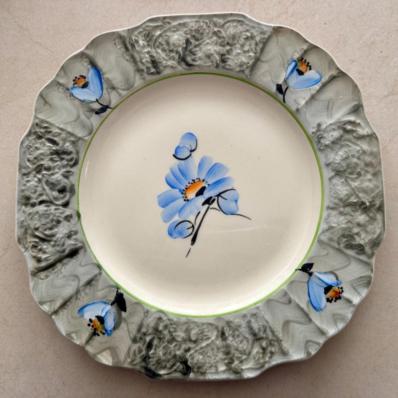 Op de afbeelding: Een ronde keramische bord met een geschulpte rand en een cr&egrave;mekleurig middenstuk beschilderd met een blauwe bloem. De brede rand heeft een getextureerde, gemarmerde grijze afwerking met vier handgeschilderde blauwe bloemen.