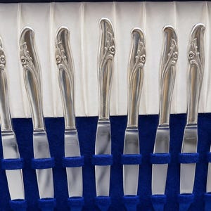 May include: A set of eight silver-toned dinner knives arranged in a row. Each knife features a polished metal blade and a decorative handle with an embossed floral motif near the top. The knives are held in place by blue fabric loops.