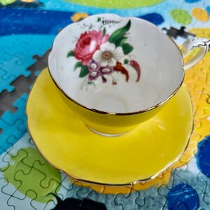 Pu&ograve; includere: Un set di tazza da t&egrave; e piattino giallo brillante con bordo smerlato e finiture dorate. L'interno della tazza presenta un'illustrazione floreale con fiori rosa, bianchi e viola e foglie verdi su sfondo bianco con un manico dorato.