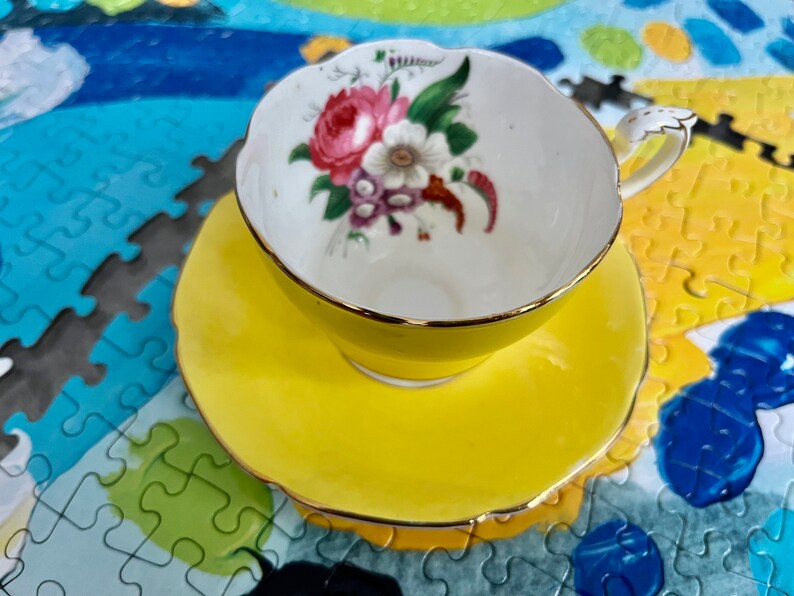 Pu&ograve; includere: Un set di tazza da t&egrave; e piattino giallo brillante con bordo smerlato e finiture dorate. L'interno della tazza presenta un'illustrazione floreale con fiori rosa, bianchi e viola e foglie verdi su sfondo bianco con un manico dorato.