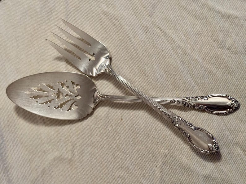K&ouml;nnte beinhalten: Ein silbernes Kuchenbesteck mit verzierten Griffen. Der Tortenheber hat ein dekoratives, durchbrochenes Design.