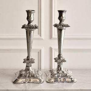 May include: A pair of silver-colored metal candlesticks featuring a tapered central column, a flared candle cup, and a square base. Each candlestick is decorated with ornate, raised floral and leaf relief patterns on the base and collar.
