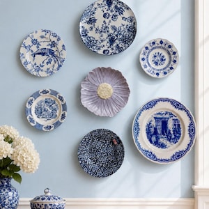 Puede incluir: Una colecci&oacute;n de seis platos decorativos de pared con varios patrones azules y blancos de flores, paisajes y aves, centrados alrededor de un plato individual en forma de flor de color lavanda con un elemento central texturizado y perlado.
