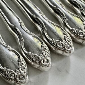 May include: Close-up of a set of antique silver spoons. The spoons have a detailed floral design on the handles and a reflective, polished surface. The spoons are arranged in a row, showcasing their intricate craftsmanship.