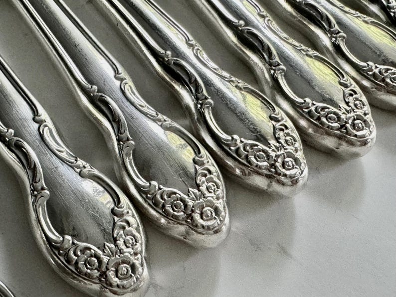 May include: Close-up of a set of antique silver spoons. The spoons have a detailed floral design on the handles and a reflective, polished surface. The spoons are arranged in a row, showcasing their intricate craftsmanship.