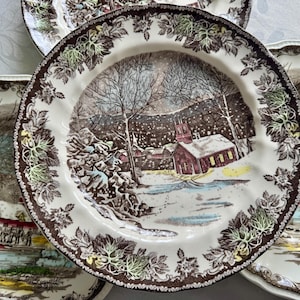 May include: Set of four brown and white ceramic dinner plates with a winter scene design. The plates feature a snowy landscape with a red schoolhouse, trees, and a river. The border of the plates is decorated with brown and green leaves.