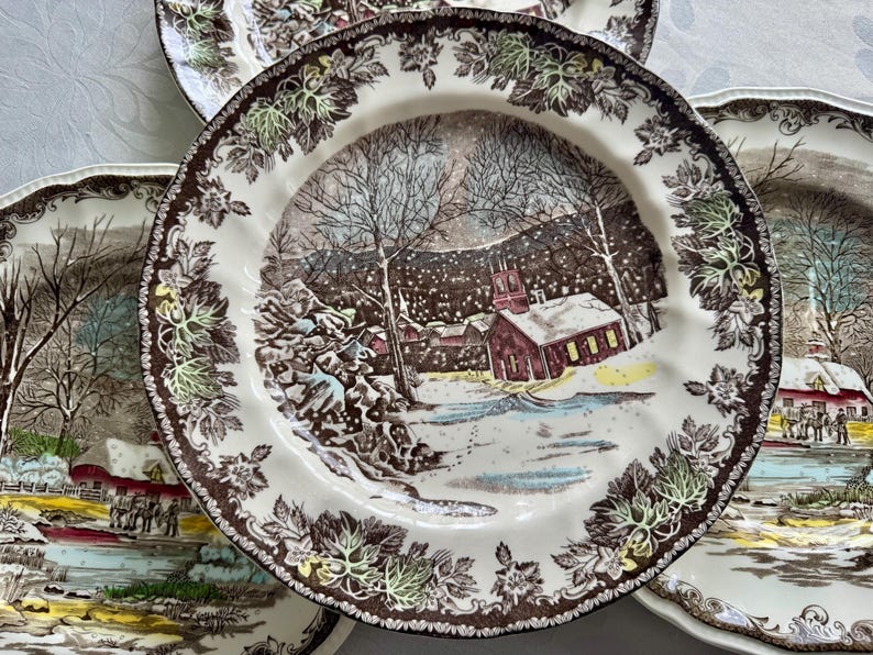 May include: Set of four brown and white ceramic dinner plates with a winter scene design. The plates feature a snowy landscape with a red schoolhouse, trees, and a river. The border of the plates is decorated with brown and green leaves.