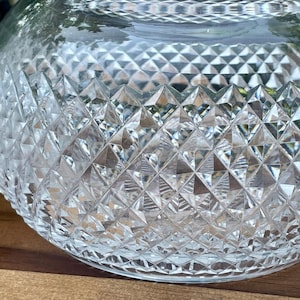 May include: A clear, round glass bowl with a diamond-cut pattern. The bowl is sitting on a wooden surface. The cut glass design creates a sparkling effect, reflecting light.
