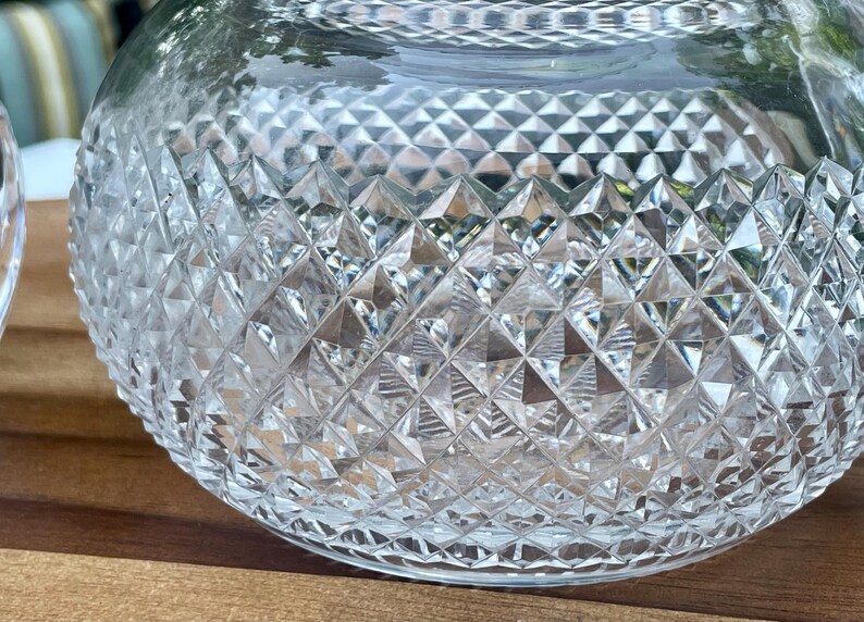 May include: A clear, round glass bowl with a diamond-cut pattern. The bowl is sitting on a wooden surface. The cut glass design creates a sparkling effect, reflecting light.
