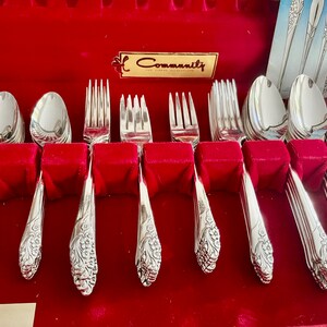 May include: A set of Community silverware in a red velvet-lined case. The set includes spoons, forks, and knives with ornate floral detailing. The case has a gold label that reads "Community".