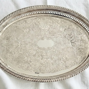 May include: An oval silver-toned serving tray featuring an intricate engraved floral and scrollwork pattern across the center, a smooth central oval area, and a decorative pierced lattice border with a twisted rope-style outer edge.
