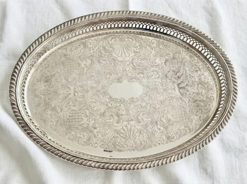 May include: An oval silver-toned serving tray featuring an intricate engraved floral and scrollwork pattern across the center, a smooth central oval area, and a decorative pierced lattice border with a twisted rope-style outer edge.