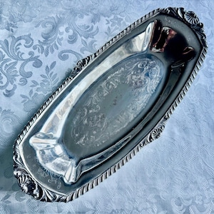 May include: A silver-toned rectangular tray with an ornate, embossed floral design. The tray has a decorative, scalloped edge and two ashtray-style indentations. The tray is likely made of metal and has a reflective surface.