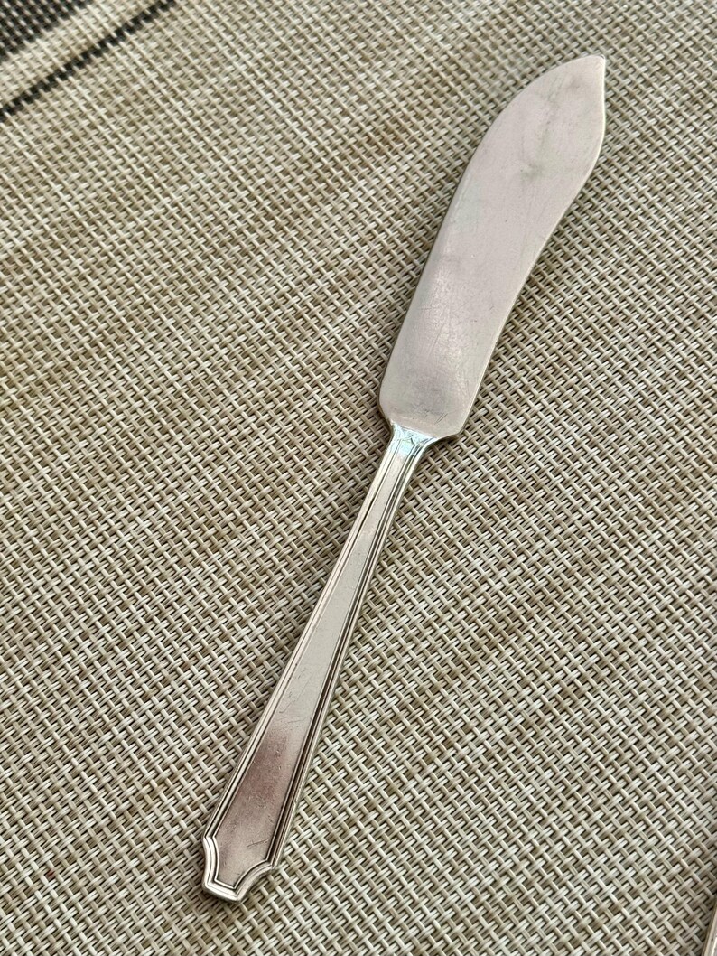 May include: A silver butter knife with a curved blade and a long, slender handle. The knife has a classic design and is made of a shiny, reflective metal. The handle has a decorative end.