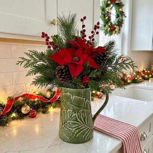 May include: A festive Christmas arrangement in a green ceramic pitcher. The arrangement includes vibrant red poinsettias, pinecones, holly, and evergreen branches. A red and white striped runner and a Christmas wreath are also visible.