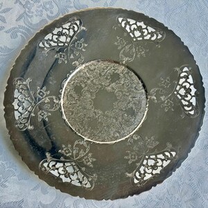 May include: A round silver-toned metal serving tray featuring a central circular area with an intricate embossed scrollwork pattern, surrounded by a smooth border with four symmetrical cut-out sections and a scalloped, decorative edge.