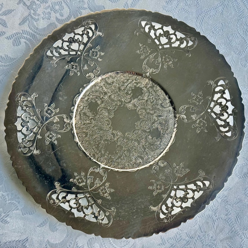 May include: A round silver-toned metal serving tray featuring a central circular area with an intricate embossed scrollwork pattern, surrounded by a smooth border with four symmetrical cut-out sections and a scalloped, decorative edge.