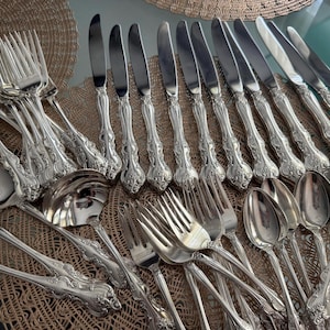 May include: An assortment of ornate silver-colored flatware, including forks, knives, and spoons, arranged on a woven placemat. The silverware features detailed floral patterns on the handles, reflecting light. A serving spoon and ladle are also visible.