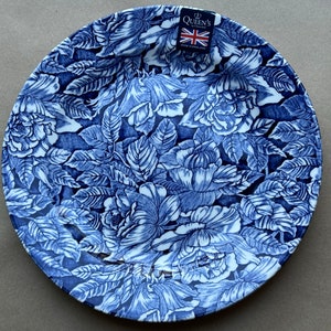 Puede incluir: Un plato redondo de cer&aacute;mica con un patr&oacute;n floral de rosas y hojas en azul oscuro y blanco. Una peque&ntilde;a etiqueta rectangular en la parte superior muestra el texto Queen's by Churchill con una bandera brit&aacute;nica y las palabras Made in England.