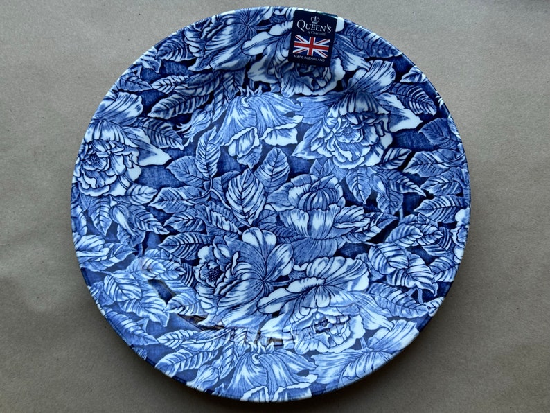 Puede incluir: Un plato redondo de cer&aacute;mica con un patr&oacute;n floral de rosas y hojas en azul oscuro y blanco. Una peque&ntilde;a etiqueta rectangular en la parte superior muestra el texto Queen's by Churchill con una bandera brit&aacute;nica y las palabras Made in England.