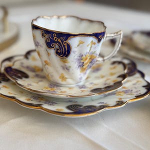 May include: A vintage teacup and saucer set with a white base, blue and gold floral detailing, and a scalloped edge. The teacup sits on a matching saucer. The set is part of a tea service, with a delicate handle.