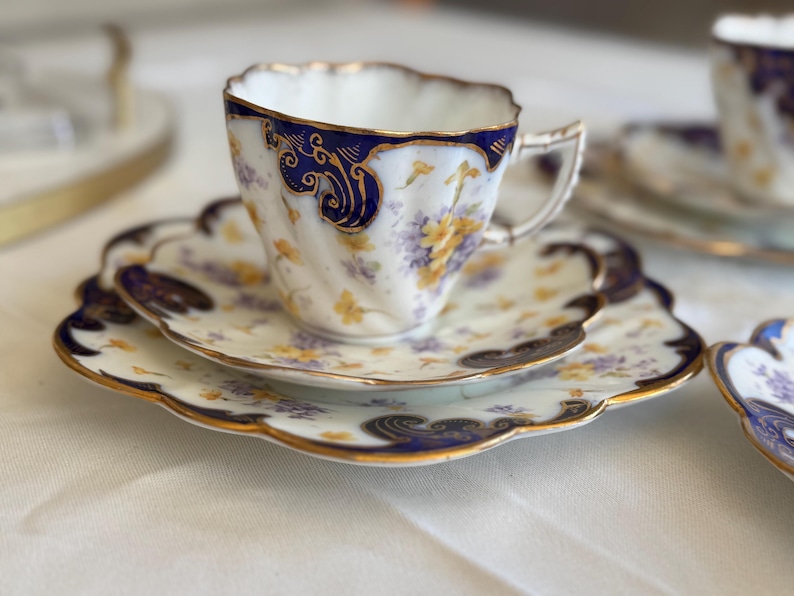 May include: A vintage teacup and saucer set with a white base, blue and gold floral detailing, and a scalloped edge. The teacup sits on a matching saucer. The set is part of a tea service, with a delicate handle.