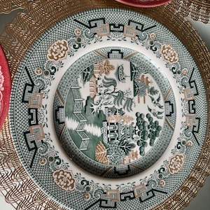 May include: A white and green decorative plate with a detailed landscape design. The plate features a green border with geometric patterns and floral accents. A gold-colored placemat is visible.