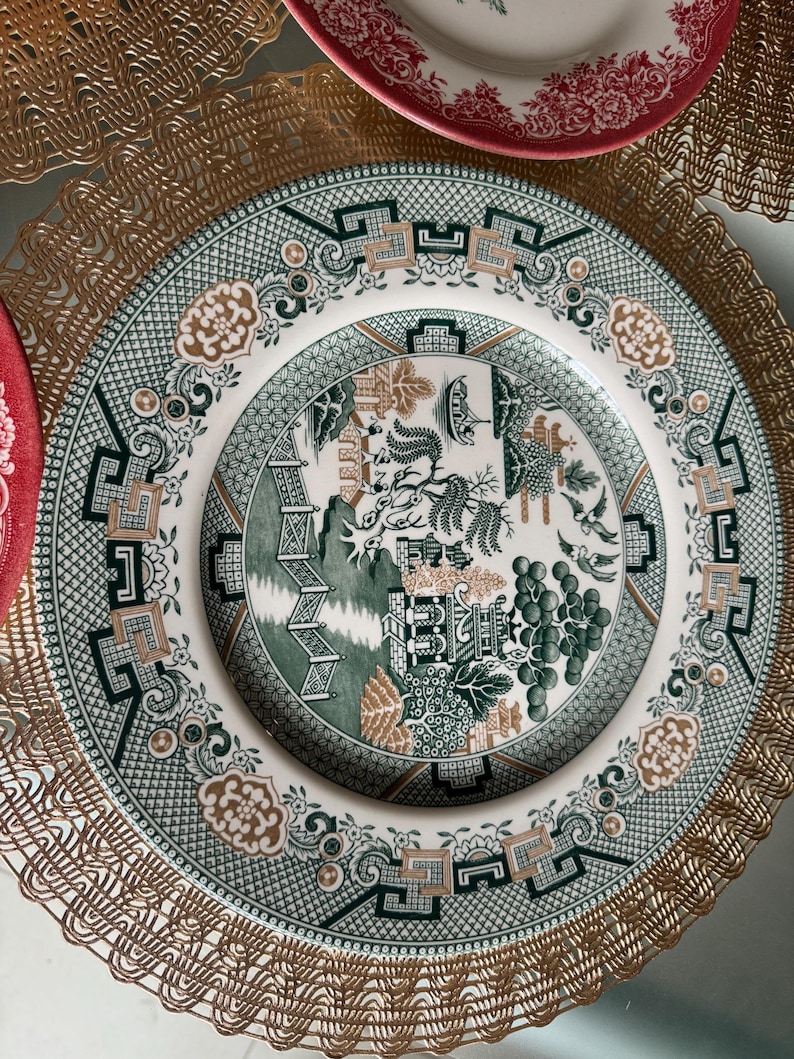 May include: A white and green decorative plate with a detailed landscape design. The plate features a green border with geometric patterns and floral accents. A gold-colored placemat is visible.