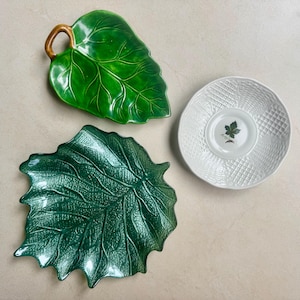 Puede incluir: Dos platos de cer&aacute;mica con forma de hojas verdes con venas detalladas y un asa de tallo marr&oacute;n, junto a un platillo circular blanco con un patr&oacute;n de celos&iacute;a texturizado y una peque&ntilde;a ilustraci&oacute;n de una hoja en el centro.